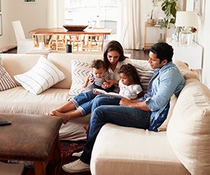 Young Hispanic family sitting on sofa reading a book together in the living room, seen from doorway