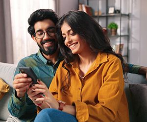 Portrait of cheerful positive young lovely couple smiling spending time together at home sitting on sofa typing on smartphone, searching internet using social network app on cellphone, family concept